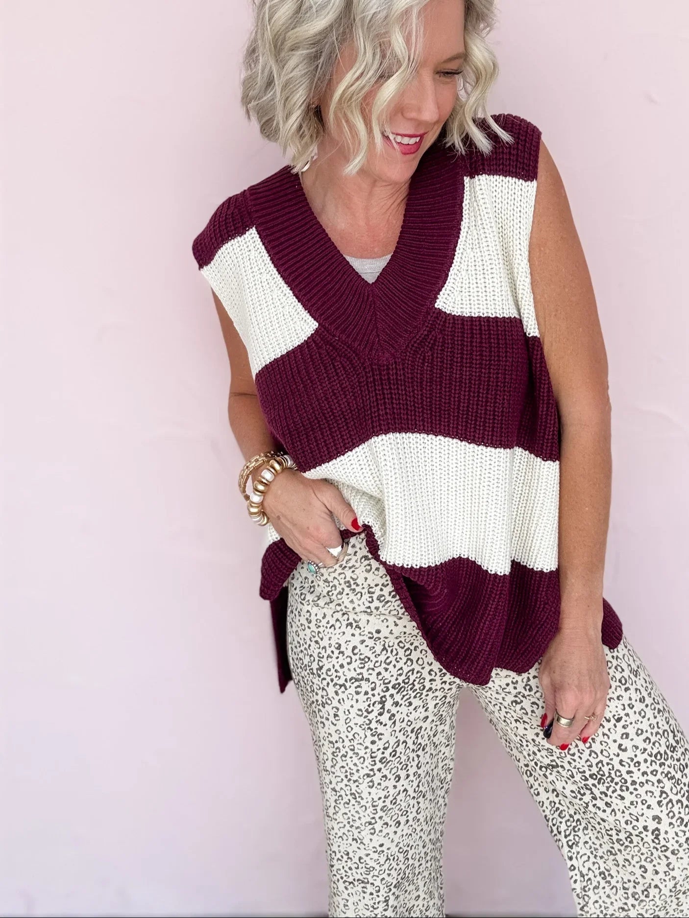 Woman wearing a sleeveless, striped sweater vest over a white top with patterned pants on a light pink background.