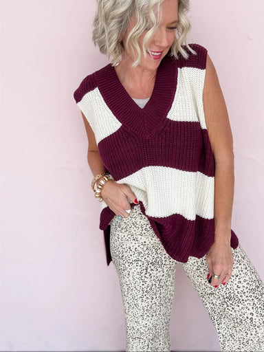 Woman wearing a sleeveless, striped sweater vest over a white top with patterned pants on a light pink background.